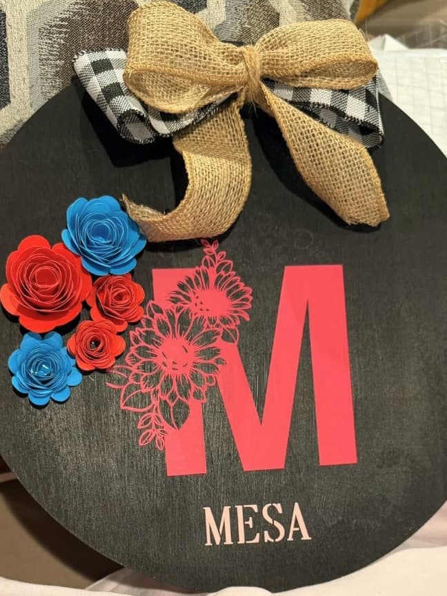 Black round door hanger with a pink M, paper flowers and a burlap bow