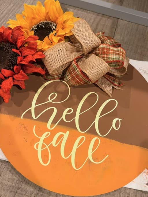 Round wood door sign reading hello fall with sunflowers and a plaid bow